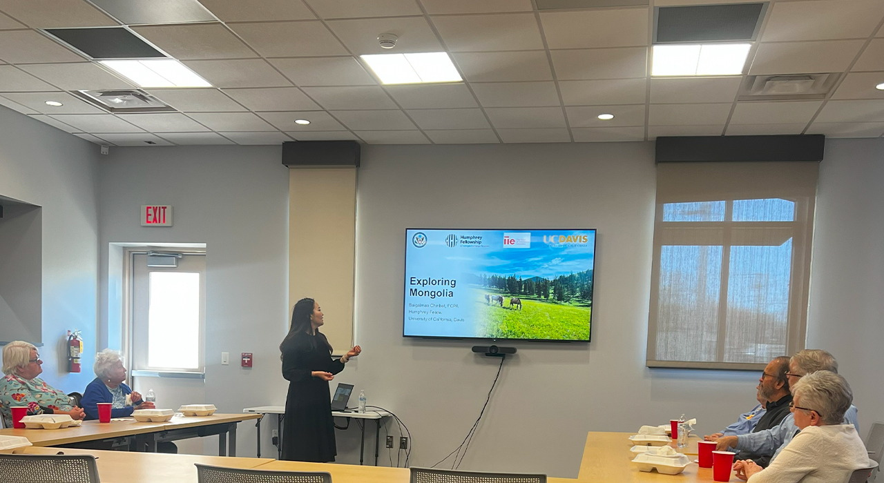 Baigalmaa stands at the front of a meeting room near a large monitor with a presentation title slide that reads "Exploring Mongolia." She gestures to the screen as attendees seated at tables surrounding her watch. On the tables are takeaway containers and red plastic cups, suggesting a working lunch meeting.