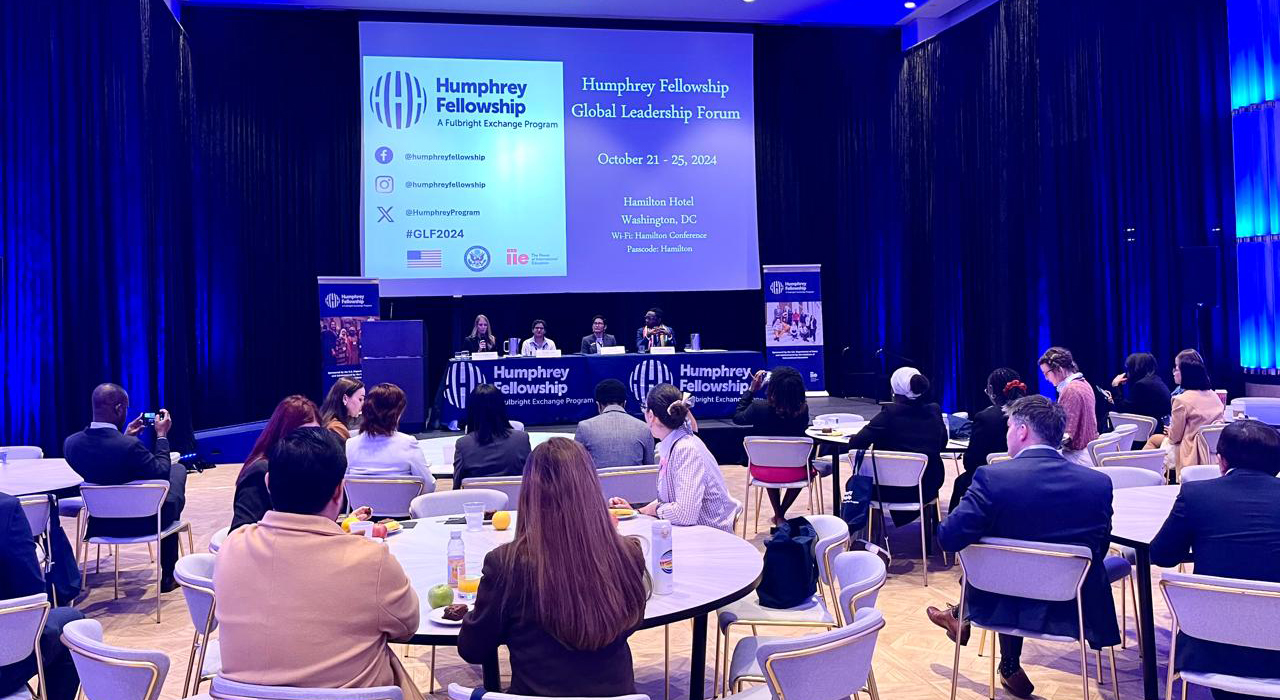 In a large conference room draped in blue, attendees sit around several round tables and focus on the panel of three speakers and a moderator at the front of the room seated at a table on a riser. Signage indicates this is the Humphrey Fellowship Global Leadership Forum 2024.