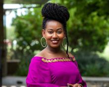 Margret wears a traditional Ugandan off-the-shoulder dress in fuchsia with gold embellishments across the top. She wears large hoop earrings and her braids are styled on the top of her head.