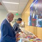 Gary May wears a navy suit and stands in the foreground at a table against a wall as he examines a collage. In the background behind him, LeShelle May looks over the other collages filling the long table. On the wall above the table is a large collage and signage about the exhibit.