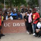 The group of 25 Fellows gathers around a short brick wall with a white UC Davis sign on it, surrounded by trees in the background.
